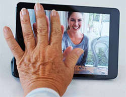 A senior man puts his hand on the screen of a tablet during a video call with a younger woman.
